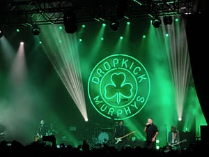 Die Dropkick Murphys auf der Bühne vor grüner Beleuchtung, einer großen grünen Neon-Vignette mit dem Bandnamen und einem Kleeblatt in der Mitte