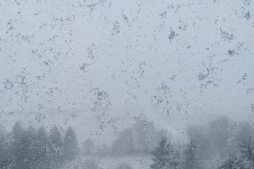 Fensterscheibe mit Schneeflocken und im Hintergrund verschwommen Felder und Bäume